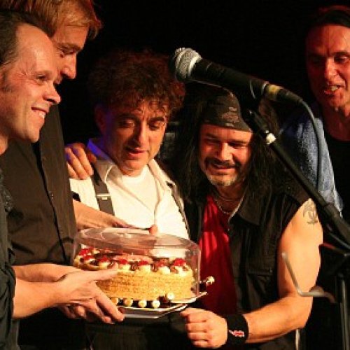 Die 5 Bandmitglieder vor dem Mikro auf der Bühne. Sie halten und schauen auf eine Torte