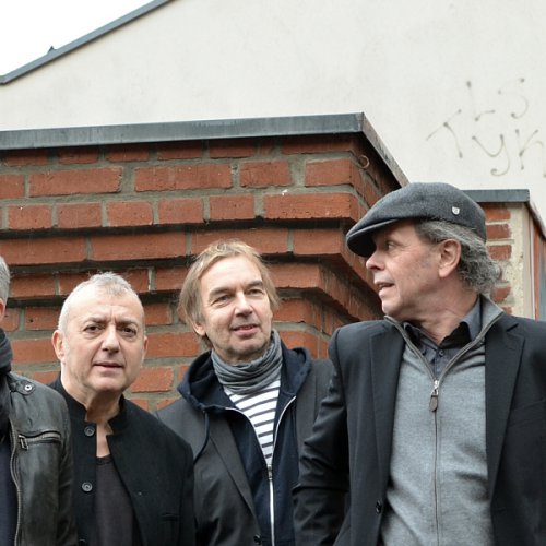 Die Band steht vor Dachaufbauten und einer Ziegelsteinwand. An der hintersten Wand rechts ein Graffiti.