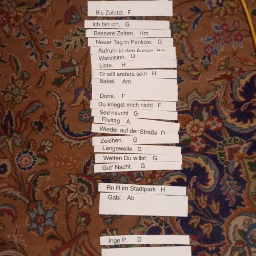 Auf einem Teppichboden liegt eine Reihe von Papierstreifen. Darauf stehen die Namen von PANKOW-Songs und die jeweilige Tonart.